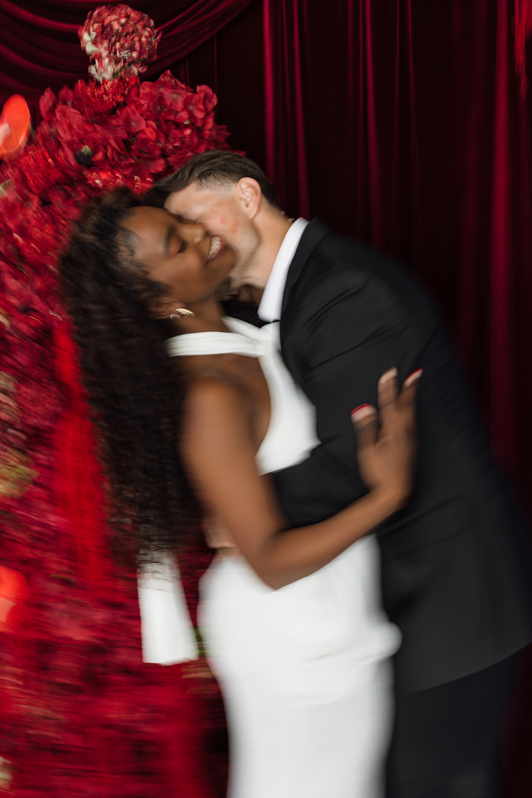 Soft, motion-blurred moment of the couple wrapped in each other’s arms, red florals filling the frame for expressive editorial engagement photos.