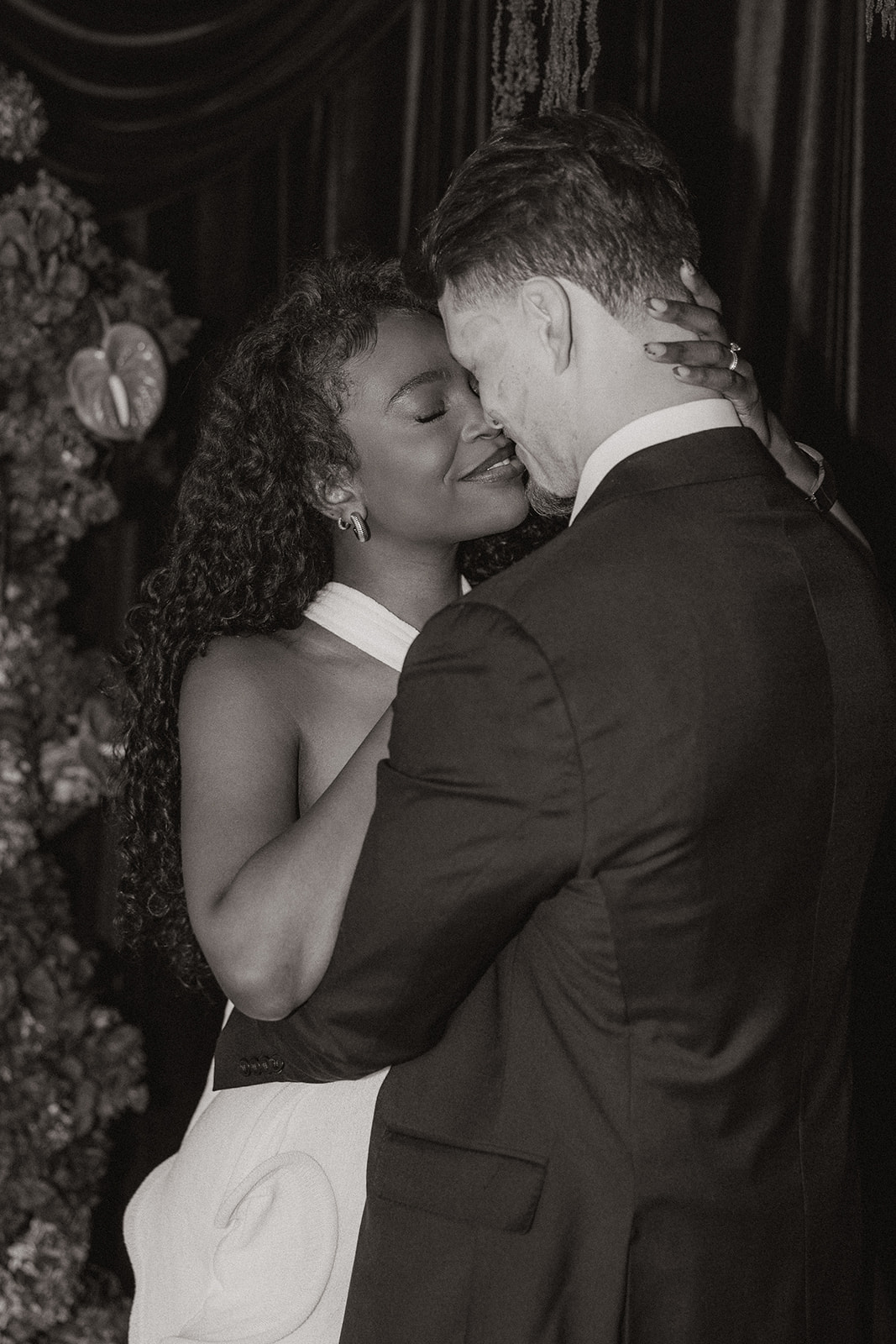 Monochrome portrait of the couple holding each other close against dark velvet drapery, eyes closed and smiling softly.