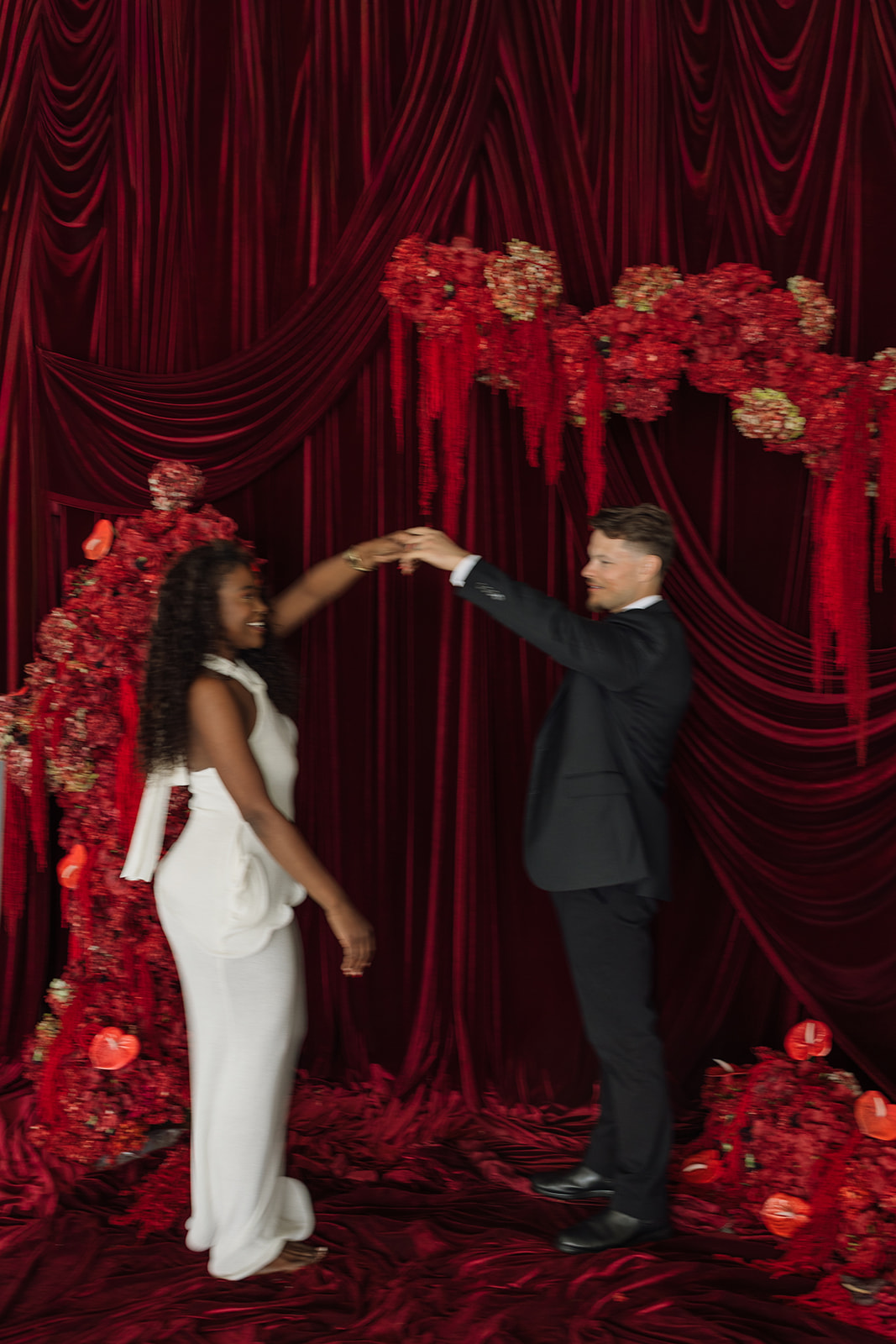 Playful twirl in front of dramatic red drapery as he spins her beneath hanging florals, blending movement and structure in editorial engagement photos.
