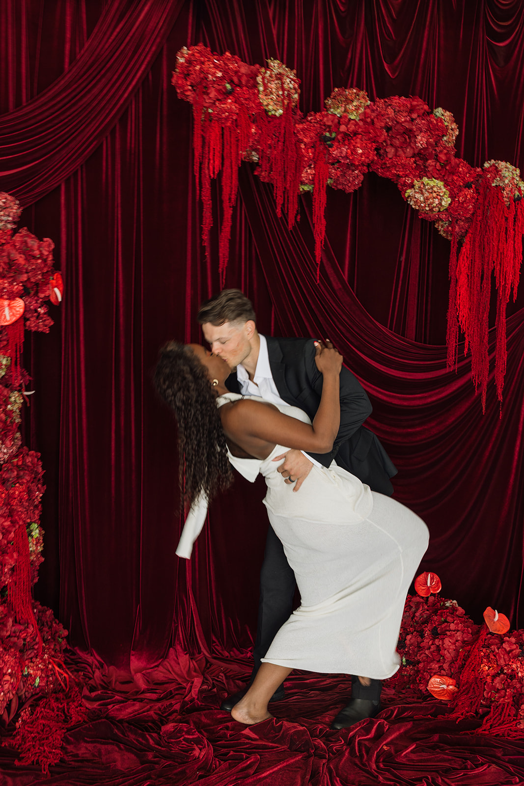 Dramatic dip kiss in front of layered velvet curtains and sculptural red floral installations, capturing movement and connection in editorial engagement photos.
