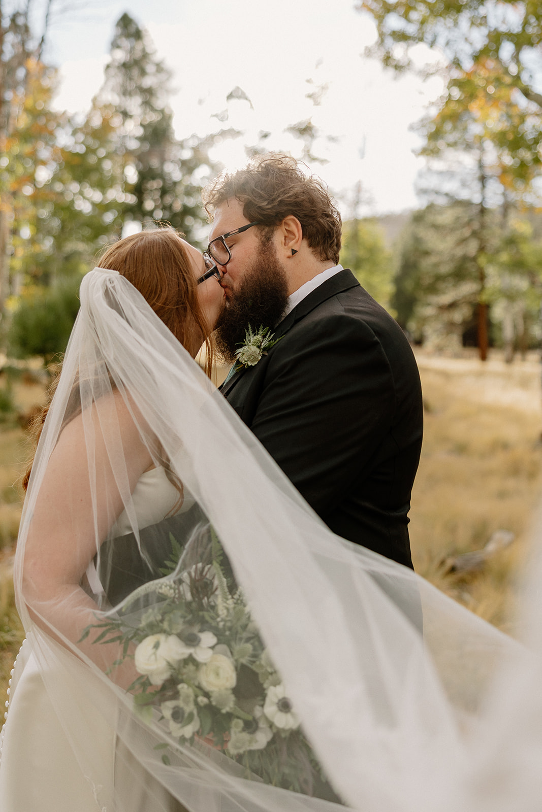Newlyweds kissing beneath a flowing veil in a wooded outdoor setting.