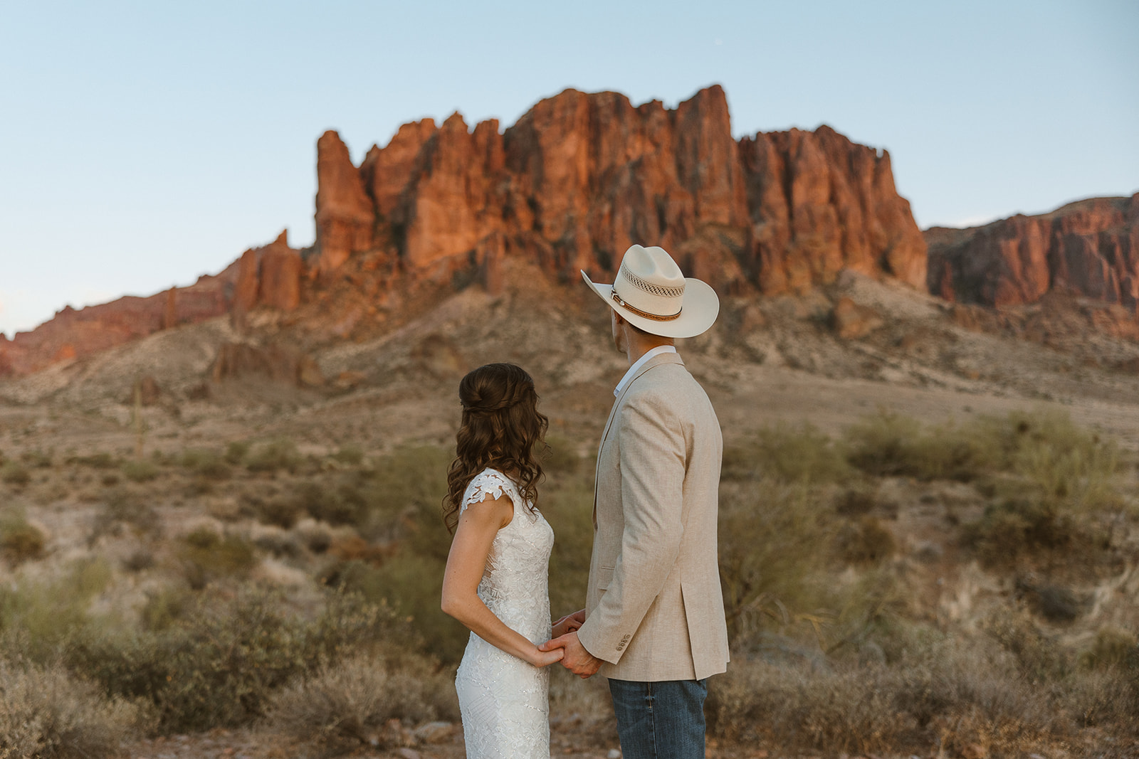 Bride and groom holding hands while facing glowing desert cliffs in the distance.