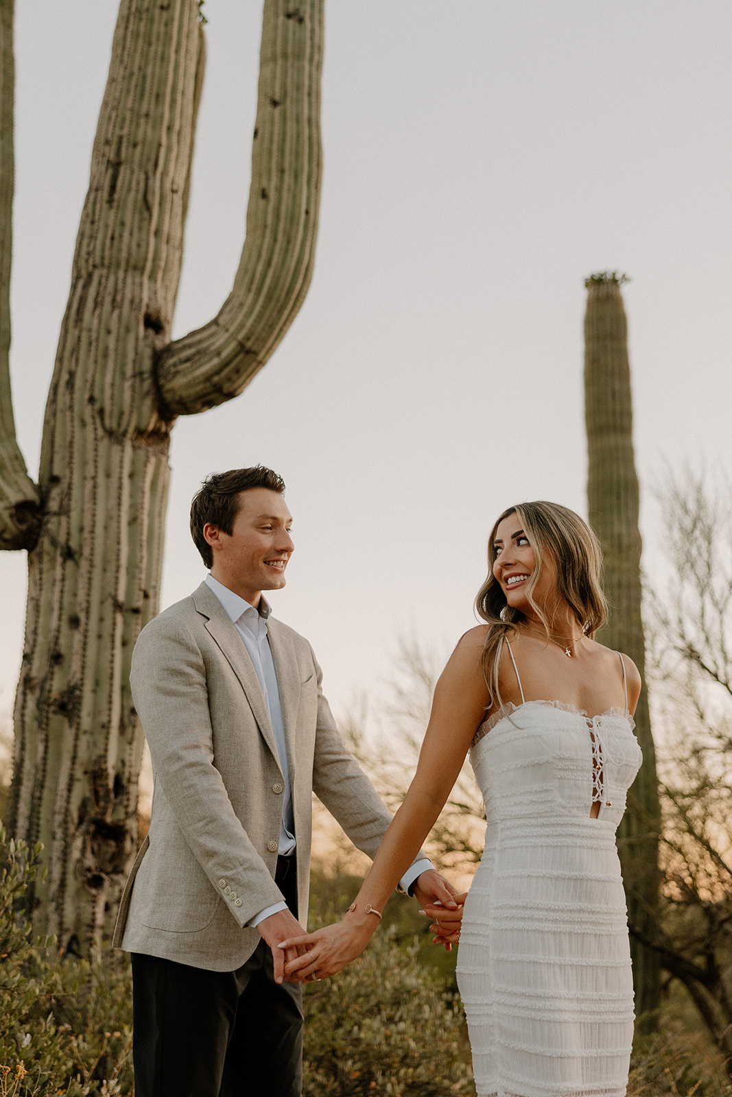 Couple smiling at each other beneath tall saguaro cacti in the Arizona desert.