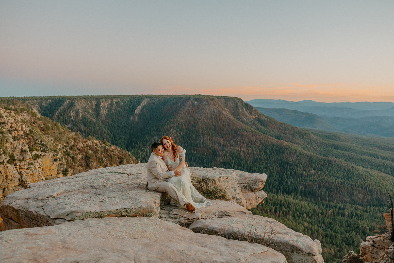 Couple sitting together on a rocky cliff overlooking layered mountain ridges at sunset.