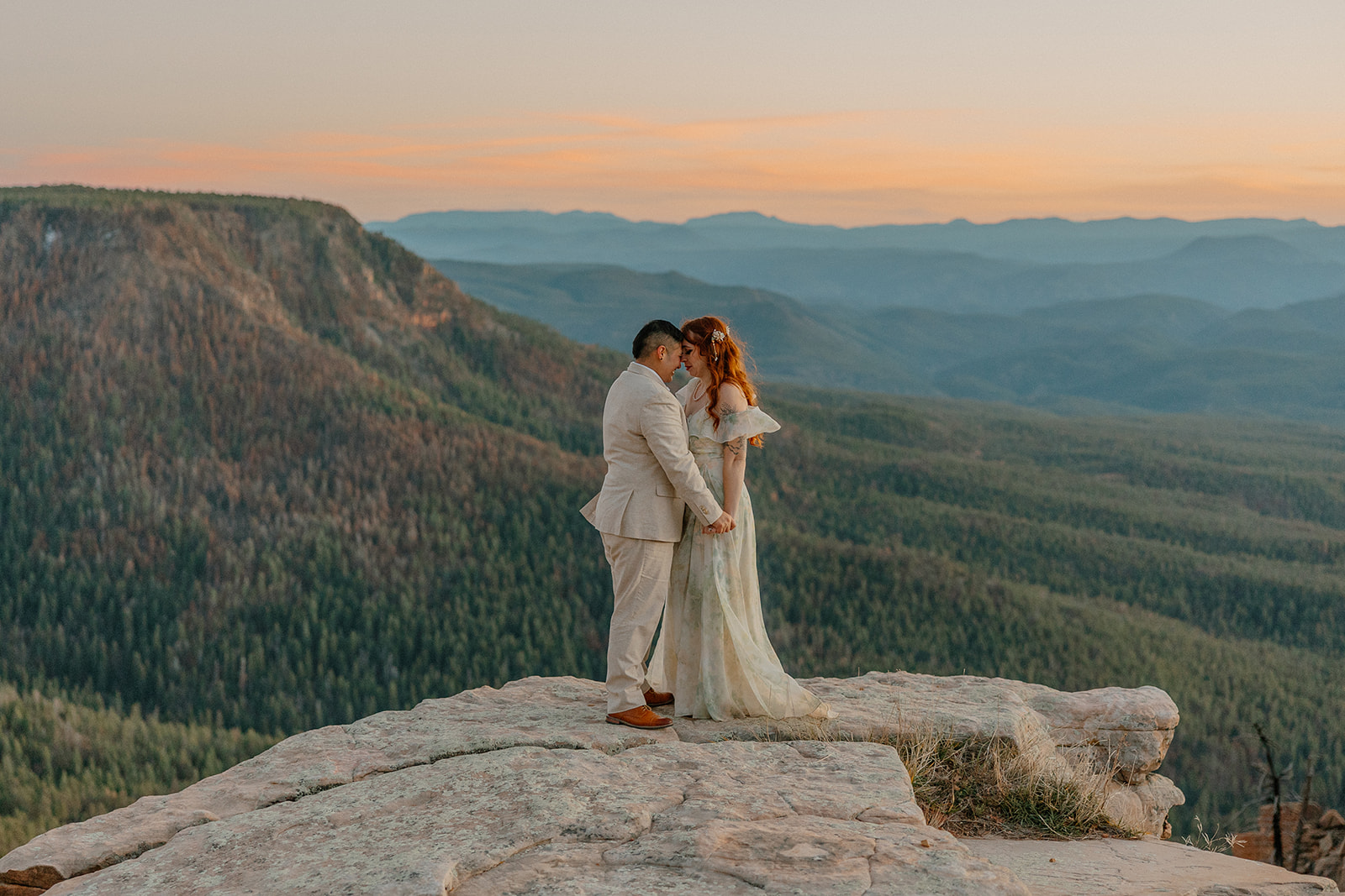 Newly married couple holding hands with foreheads touching at the side of a cliff overlooking beautiful forest. 