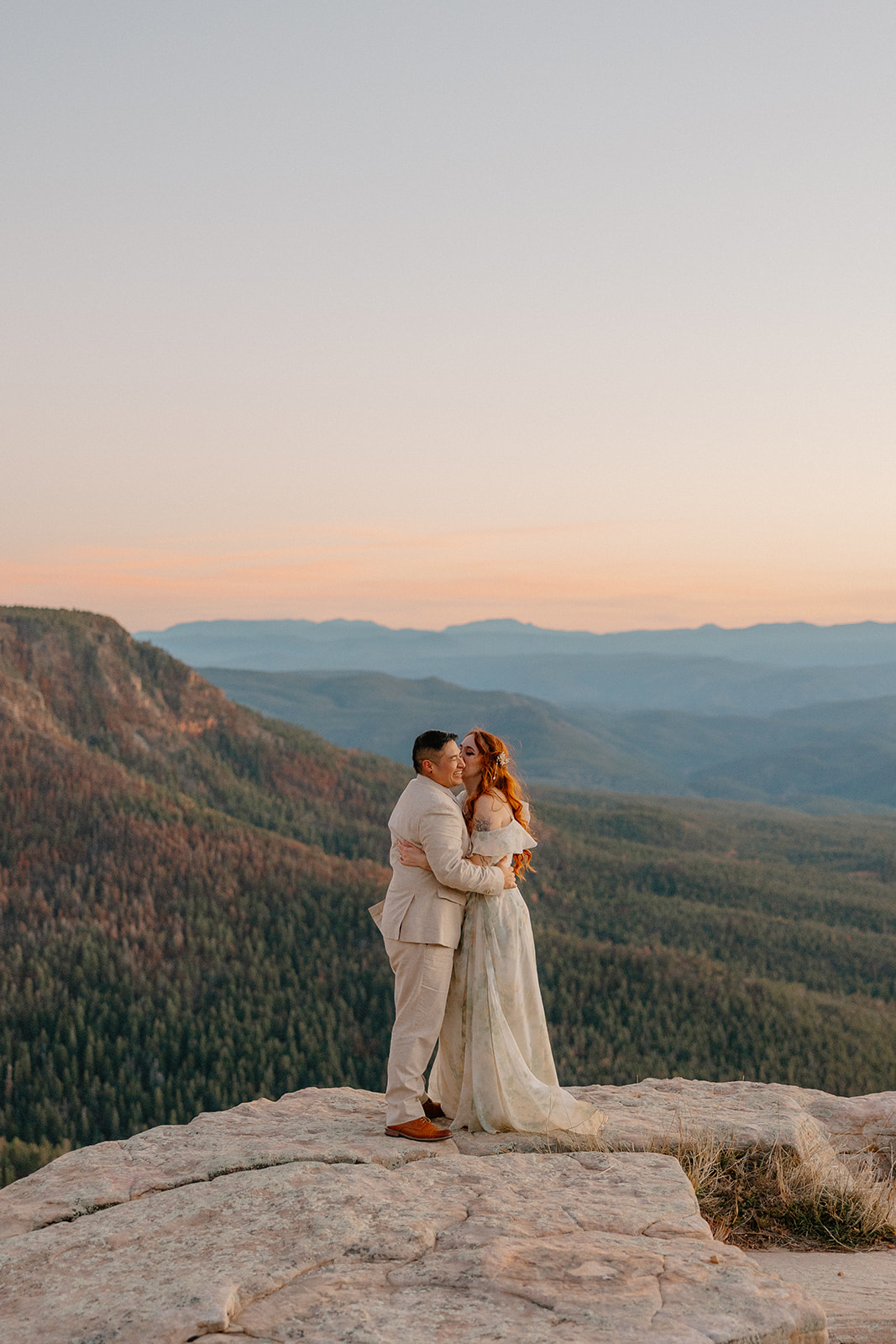 Bride and groom embracing on a cliff with endless mountain views at golden hour.