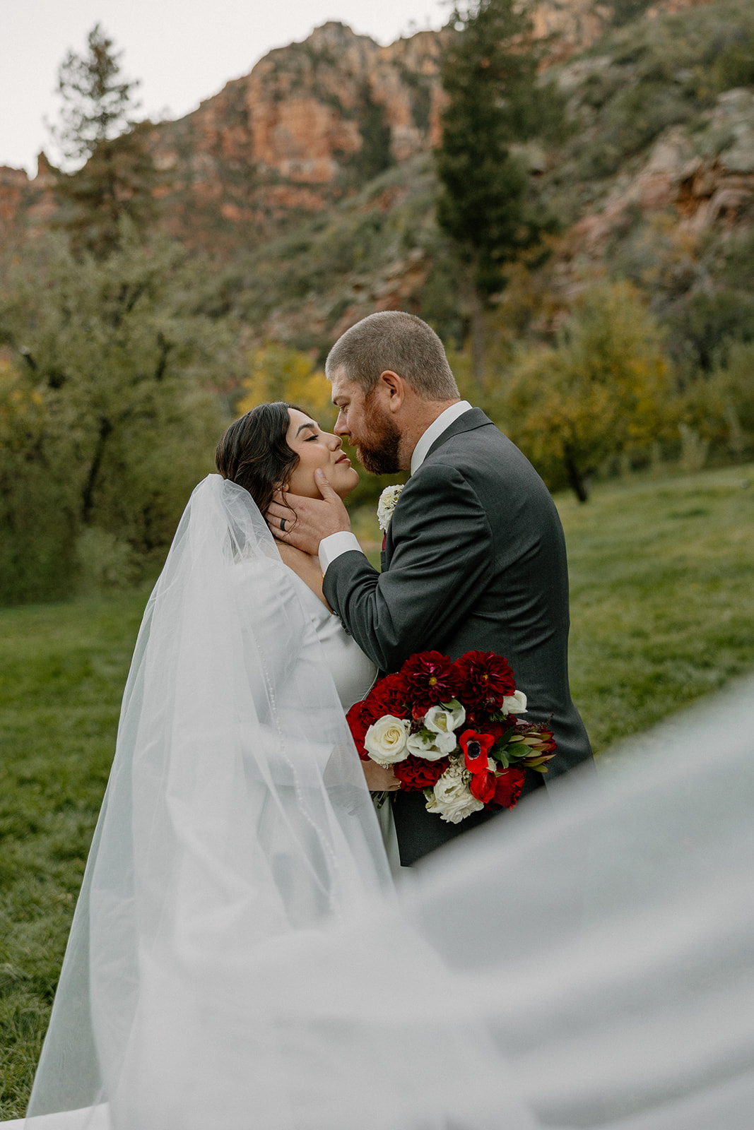 Bride and groom embracing in a green meadow with red bouquet and flowing veil.