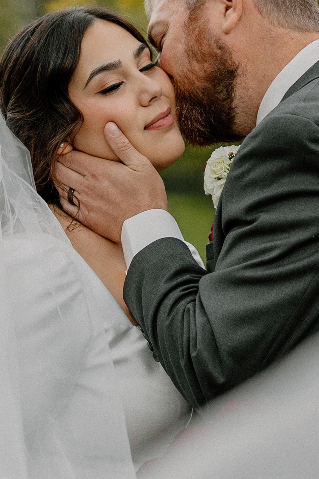 Groom holds bride's face and kisses her cheek in forested area. 