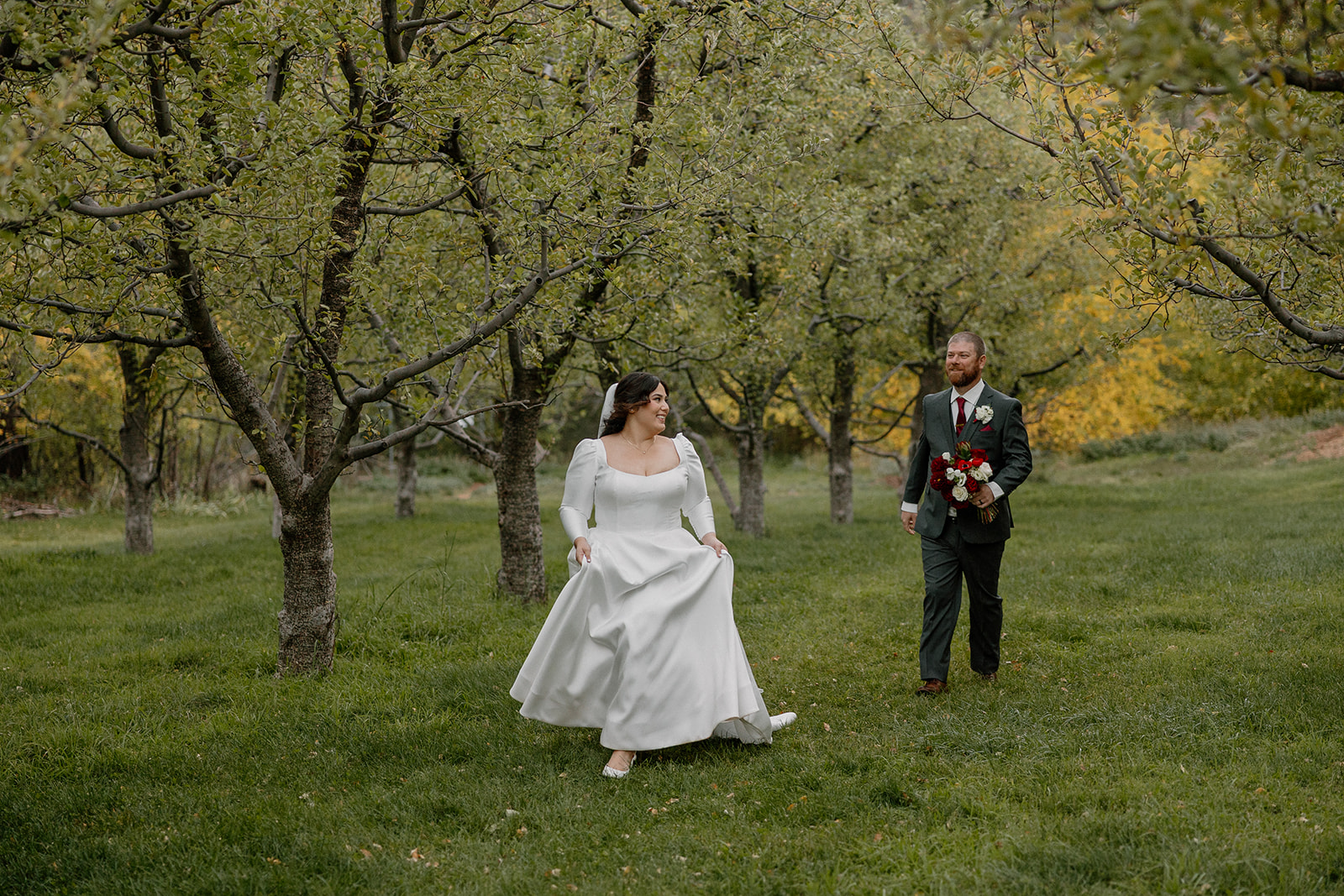 Bride walking through a green orchard toward groom holding a red bouquet.