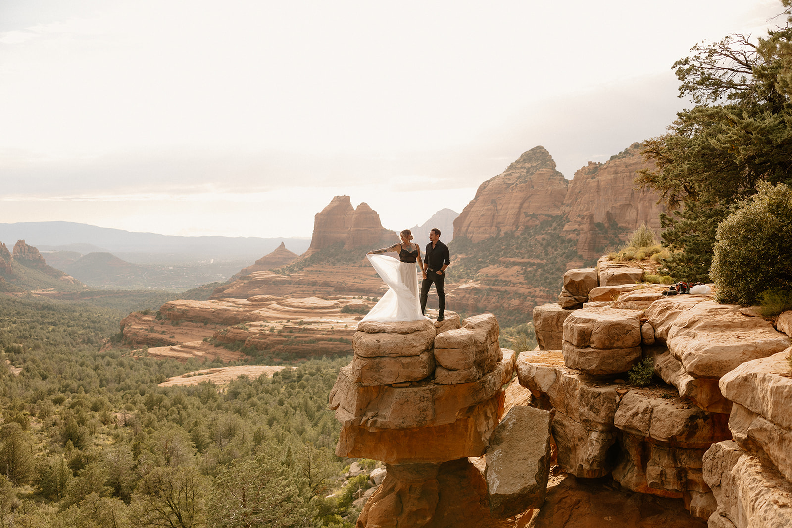 Bride lifting her dress while standing on a dramatic red rock overlook at golden hour.