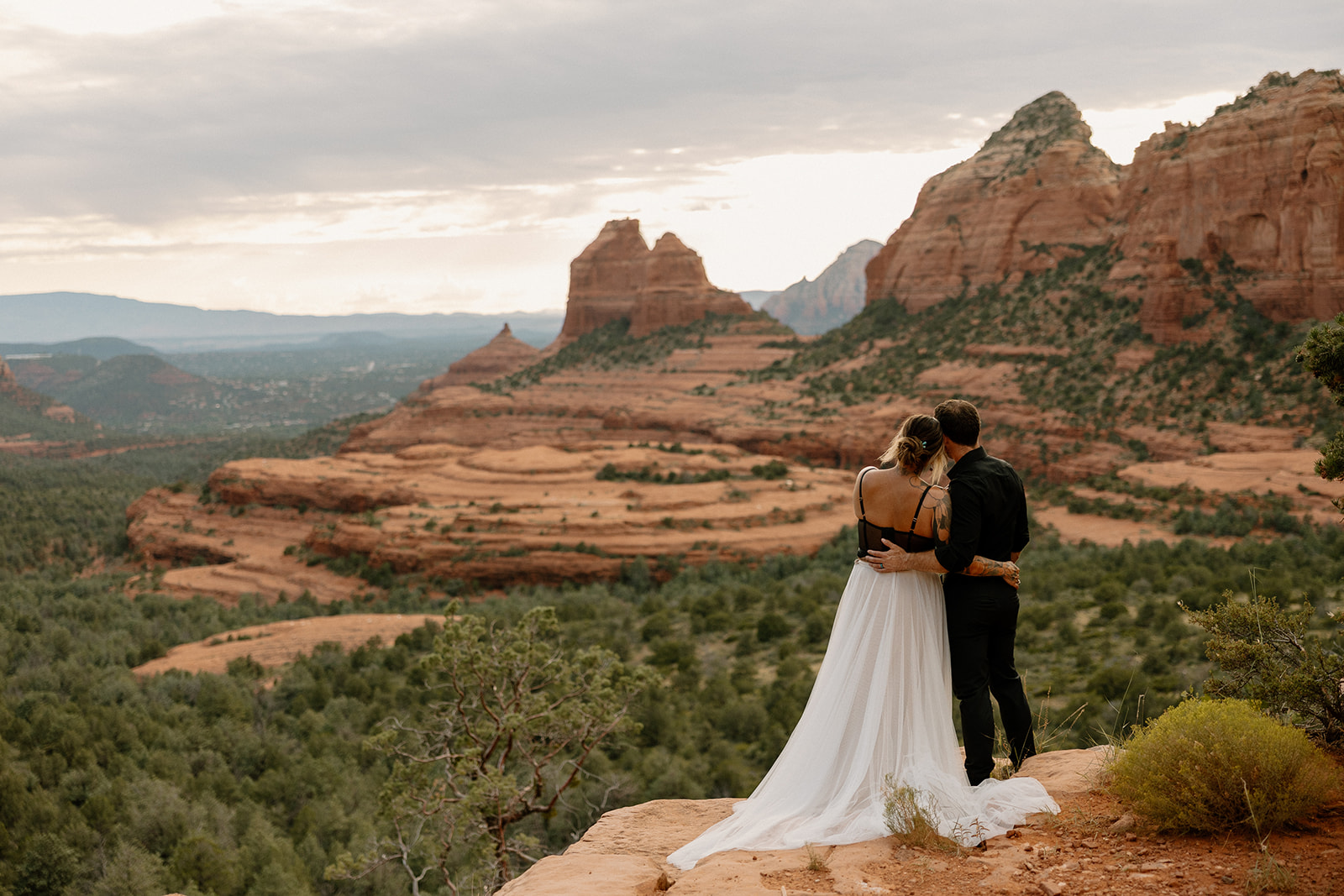 Newlyweds embracing on a cliff with sweeping red rock views at sunset.
