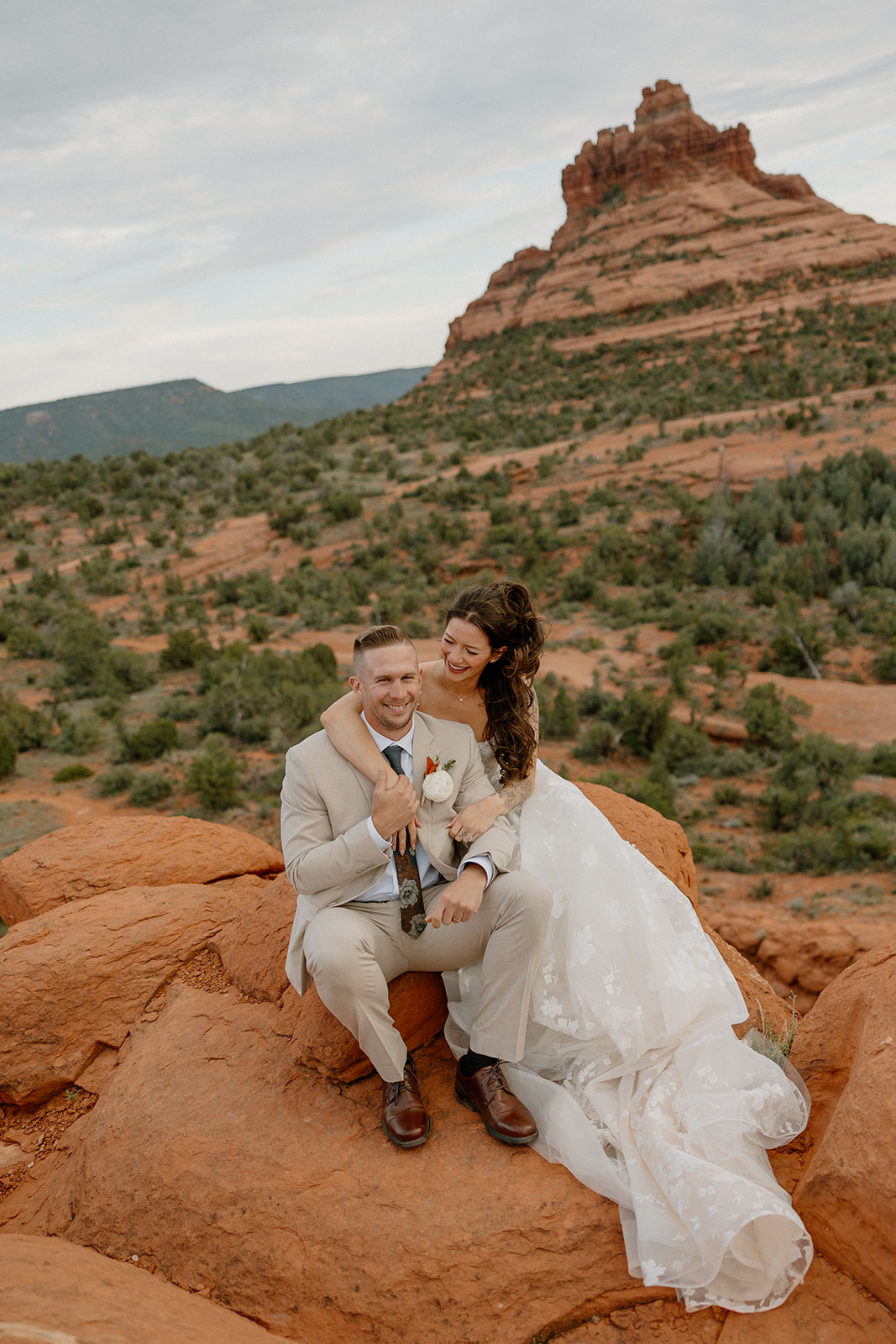 Bride and groom sitting on a red rock overlook with desert views behind them in Sedona.