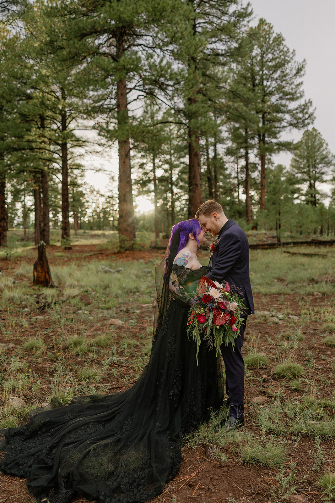 Bride in flowing black gown and groom embracing in an open pine forest meadow.