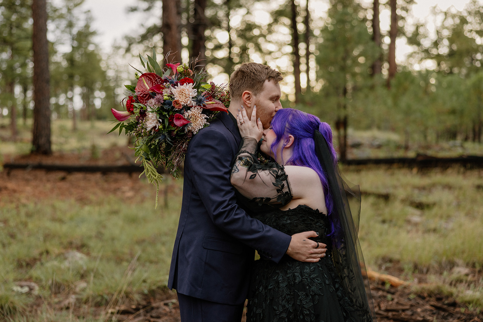 Couple sharing a kiss in a pine forest clearing with bold bouquet and black wedding attire.