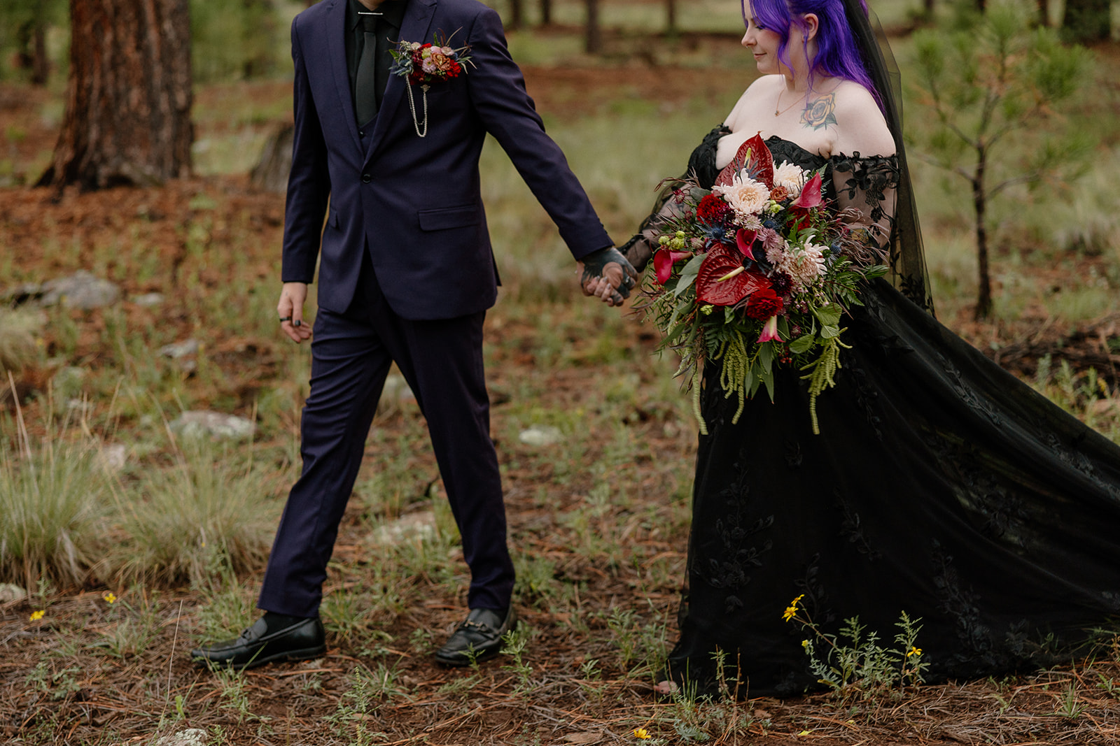 Couple in formal black wedding attire walking through a pine forest clearing.