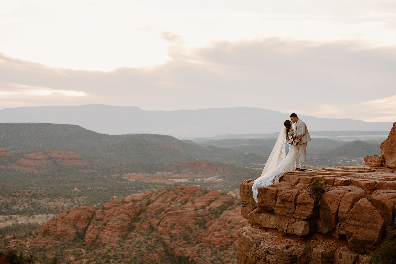 Couple standing on a cliff edge with veil blowing in the wind during their Arizona elopement.