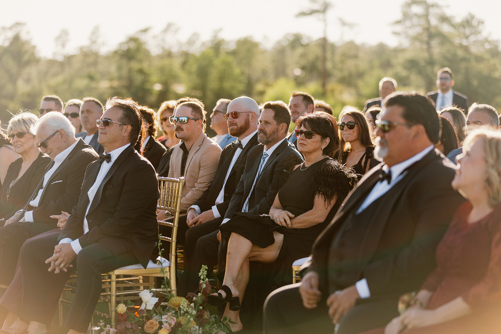 Wedding guests sit attentively during an outdoor ceremony with warm sunlight and mountain scenery.