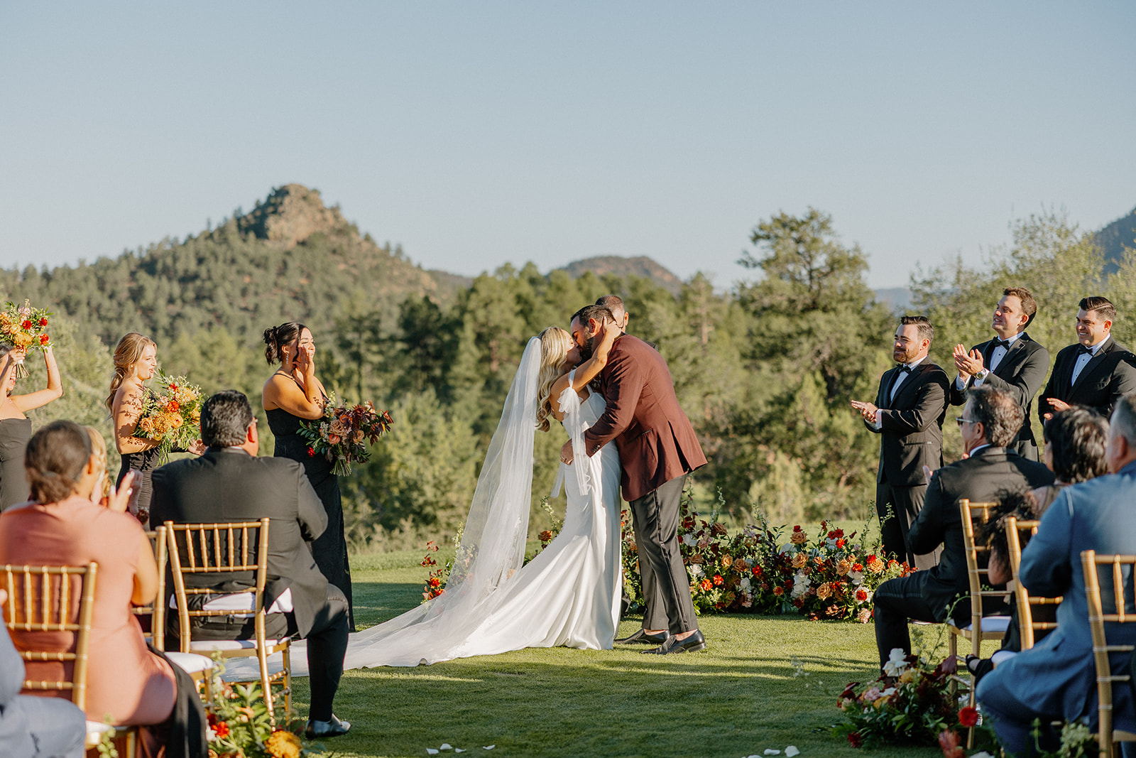 Bride and groom share their first kiss during an outdoor ceremony surrounded by florals and forest views.