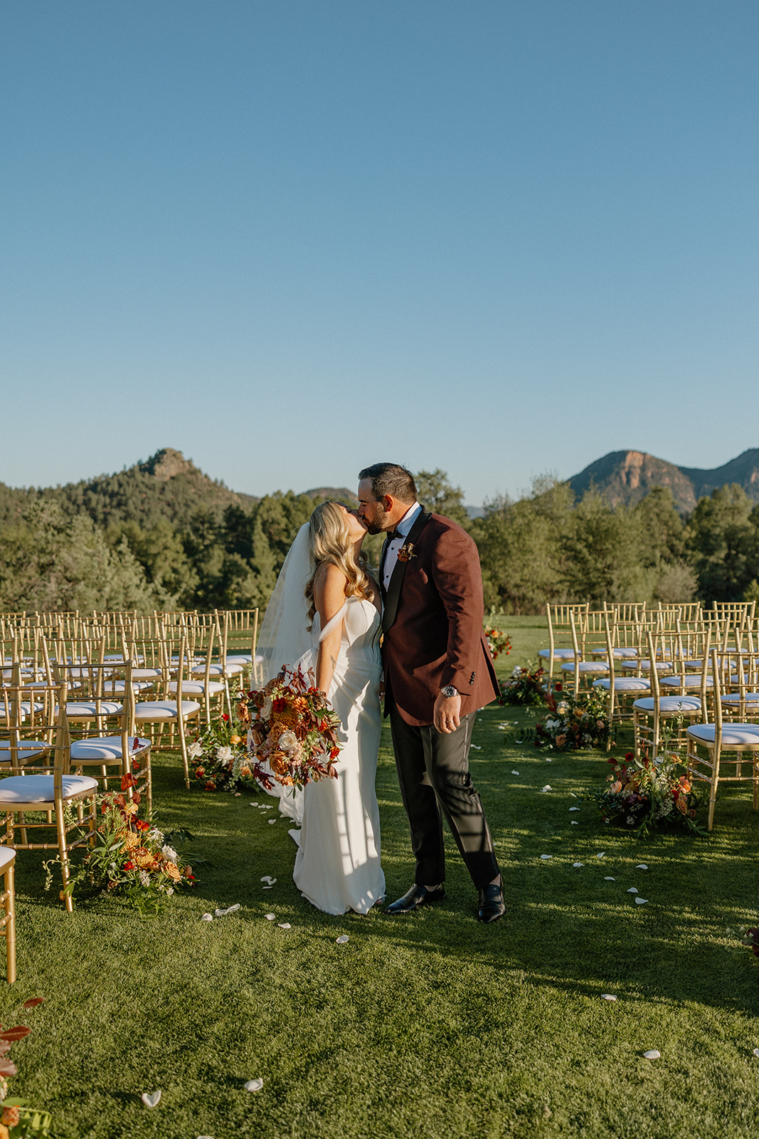 Newlyweds share a kiss in the ceremony aisle surrounded by gold chairs and florals at a Payson wedding venue.