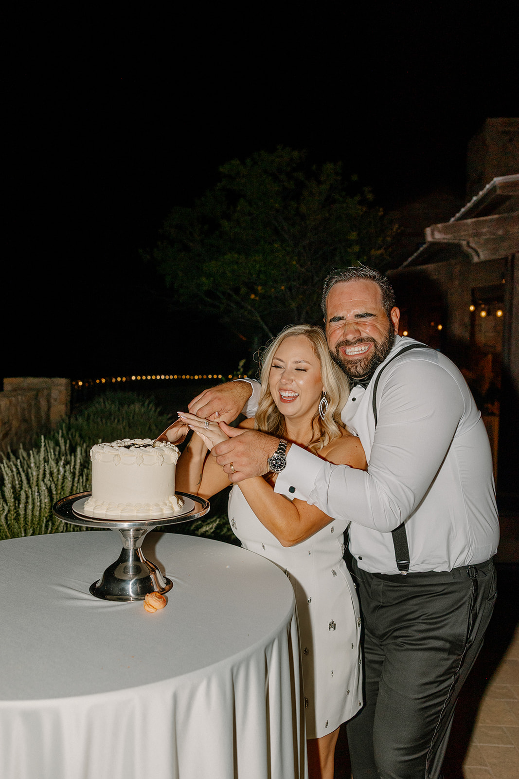 Couple laughing together while cutting their wedding cake outdoors at a Payson wedding venue under the night sky.