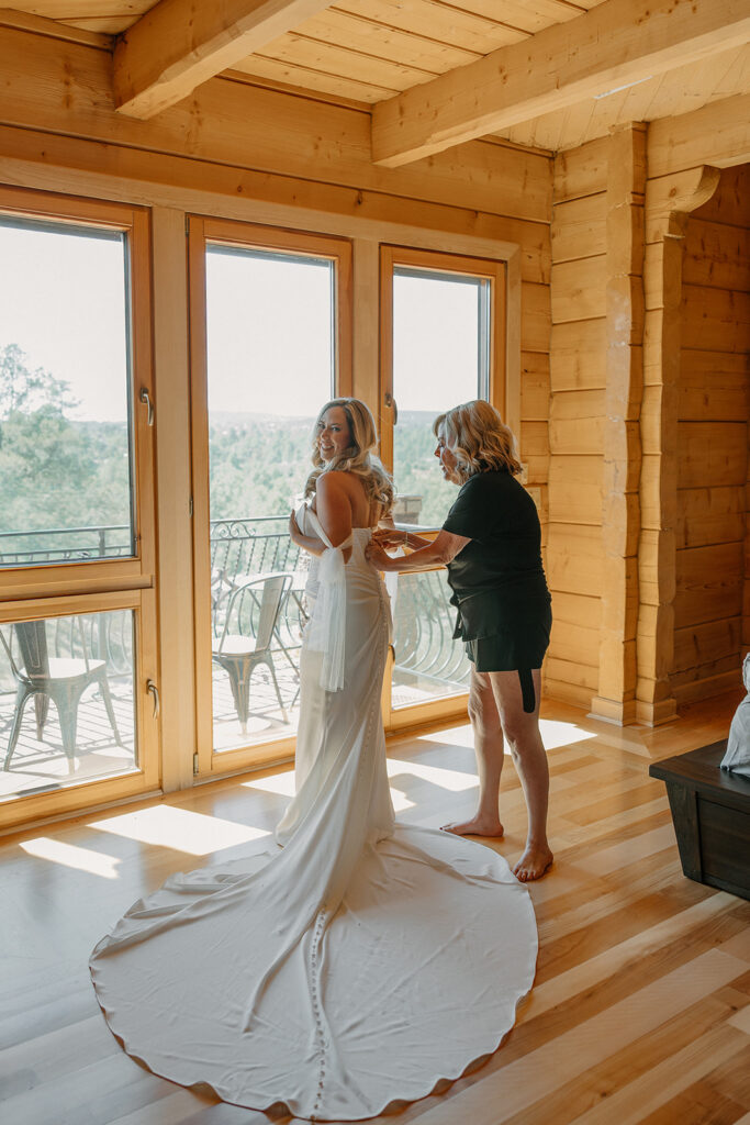 Bride stands by large windows as her dress is buttoned, light filling the room at a Payson wedding venue.