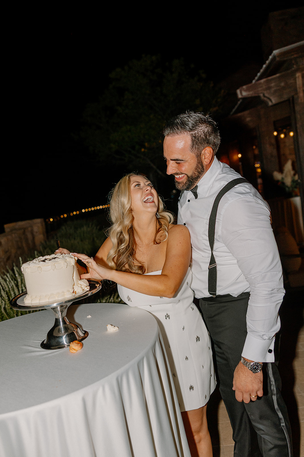 Bride and groom smile at each other while cutting their cake during their outdoor reception celebration.