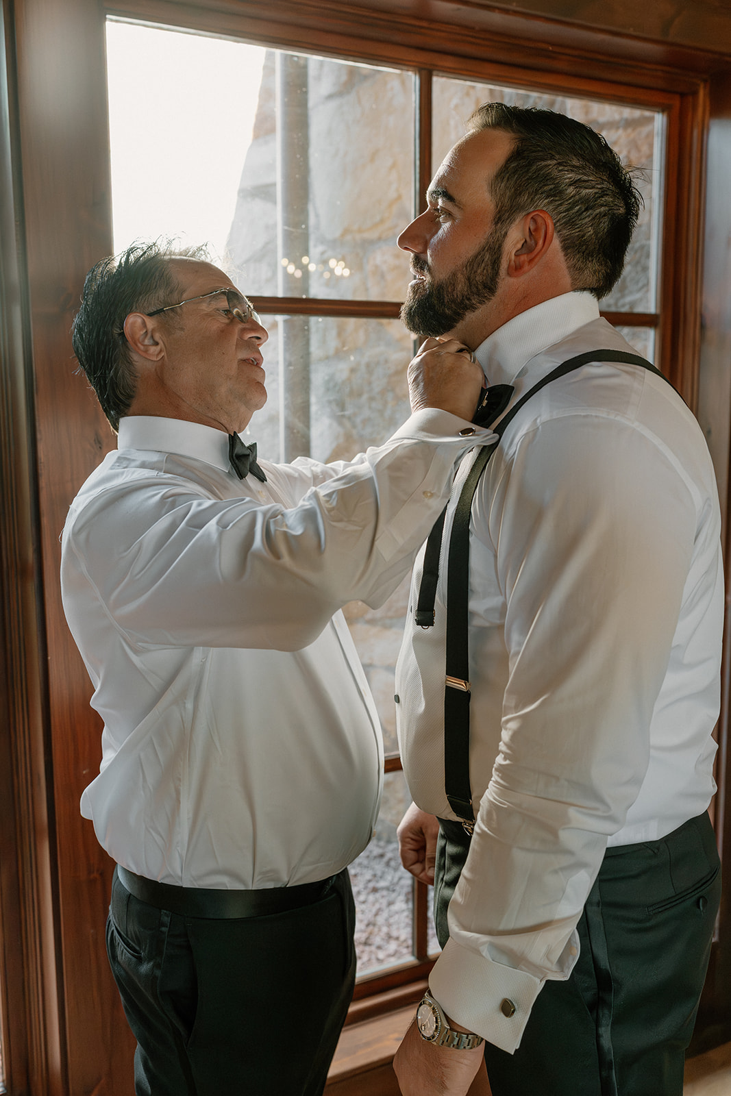 Groom’s father helps adjust his bow tie while getting ready beside a window inside a Payson wedding venue.