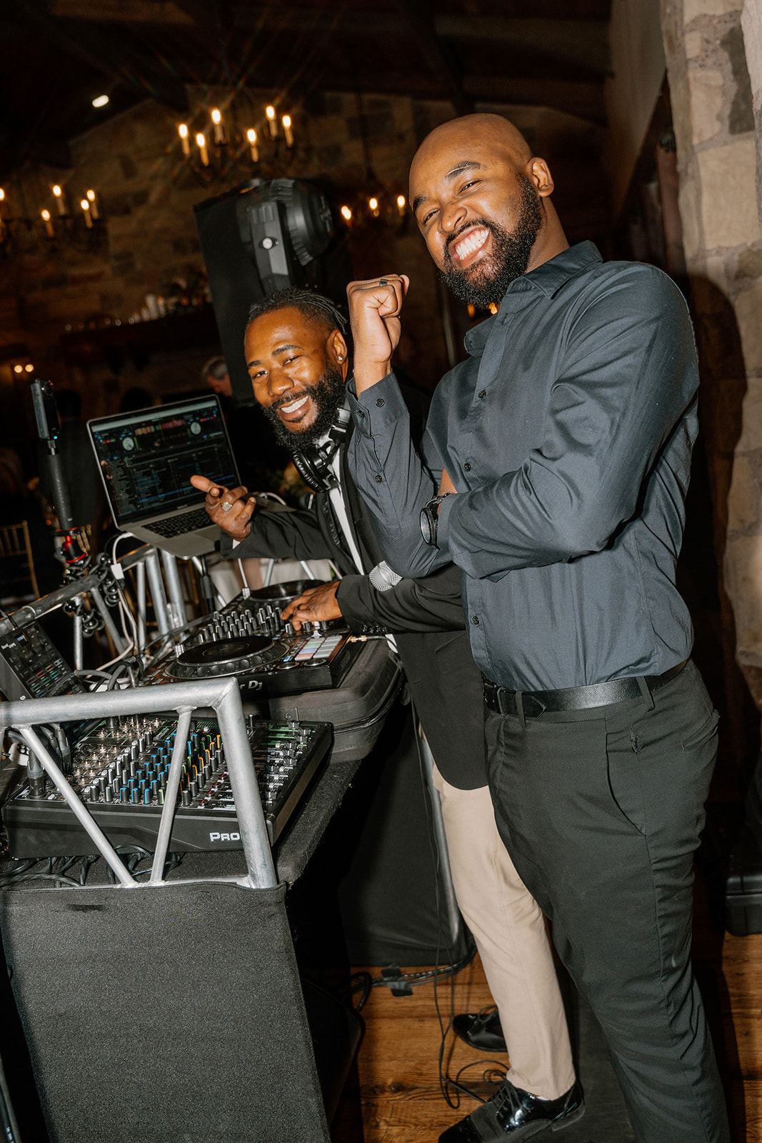 Wedding DJs smile and celebrate behind the booth while keeping the dance floor alive during a lively reception.