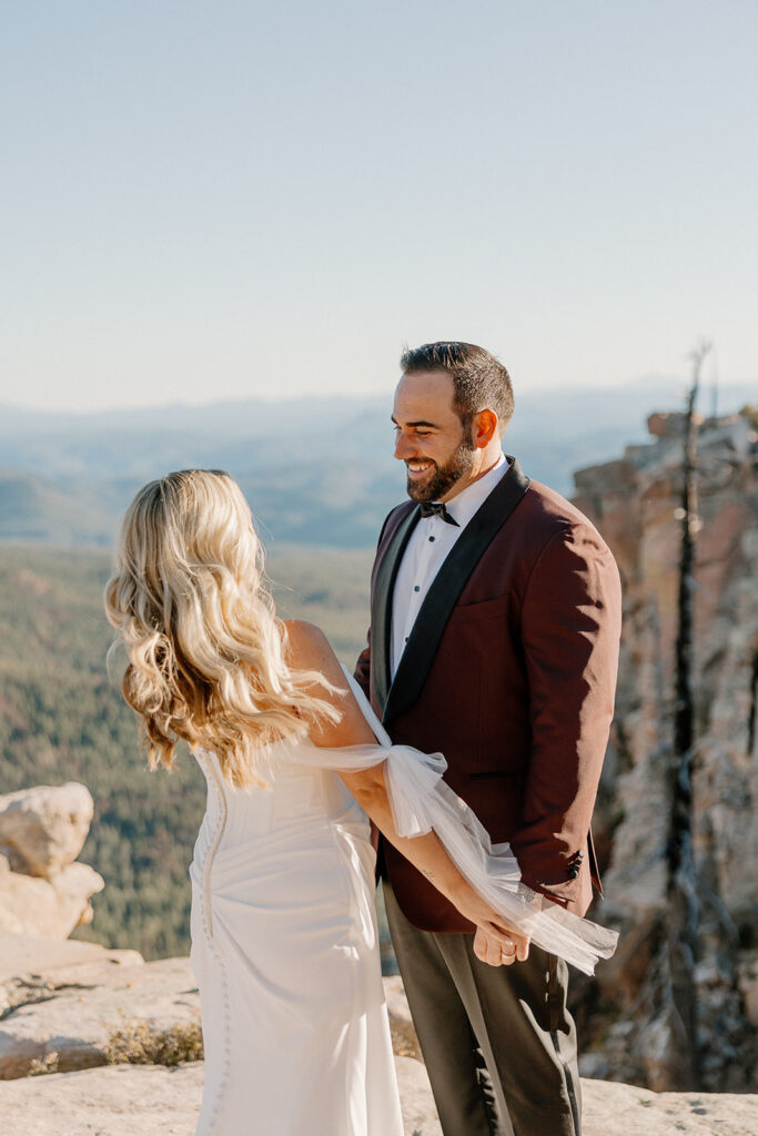 Bride and groom hold hands on a cliffside overlook, smiling at each other with sweeping forest views behind them.