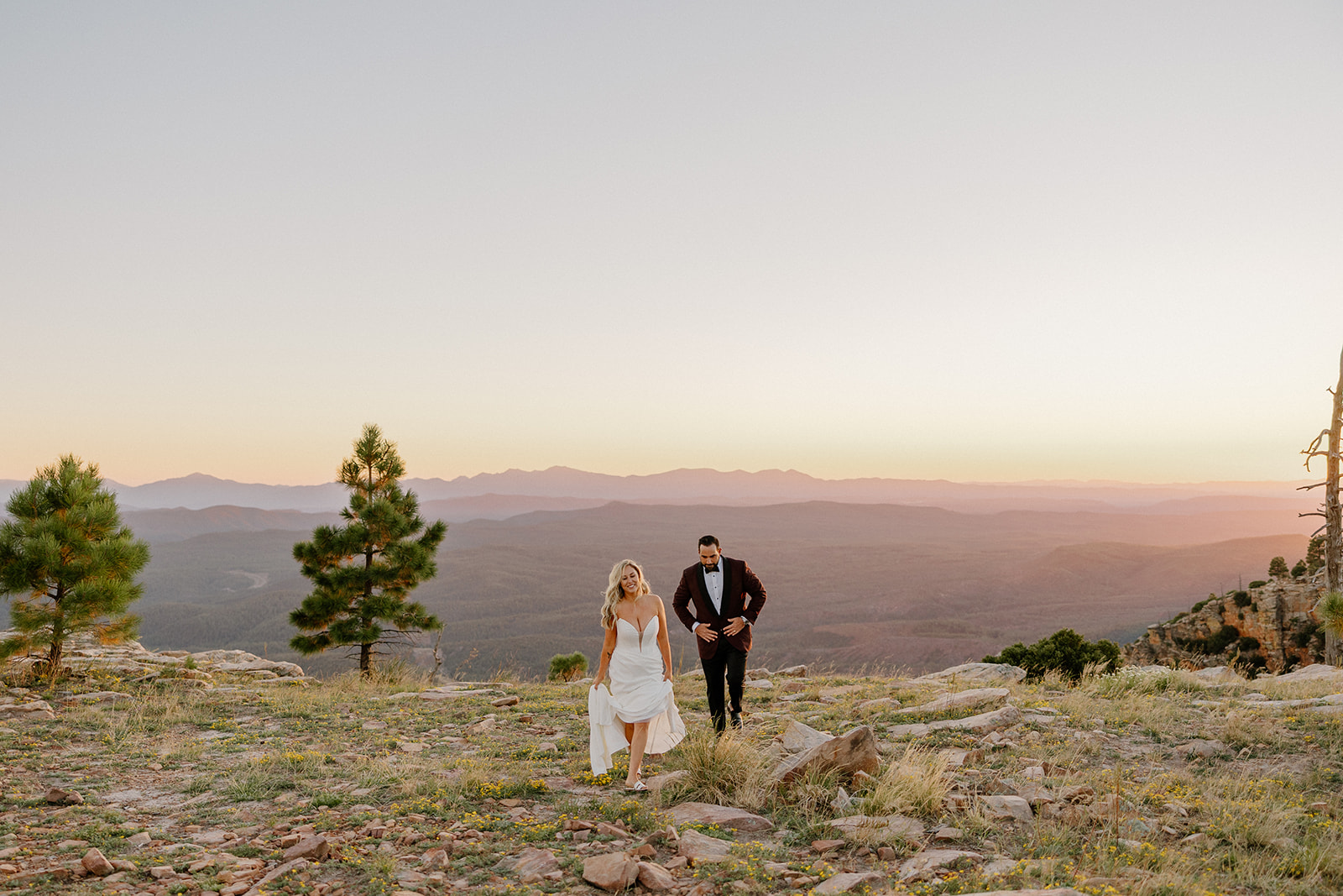 Bride and groom walk together across a rocky overlook during sunset with layered mountain views in the distance.