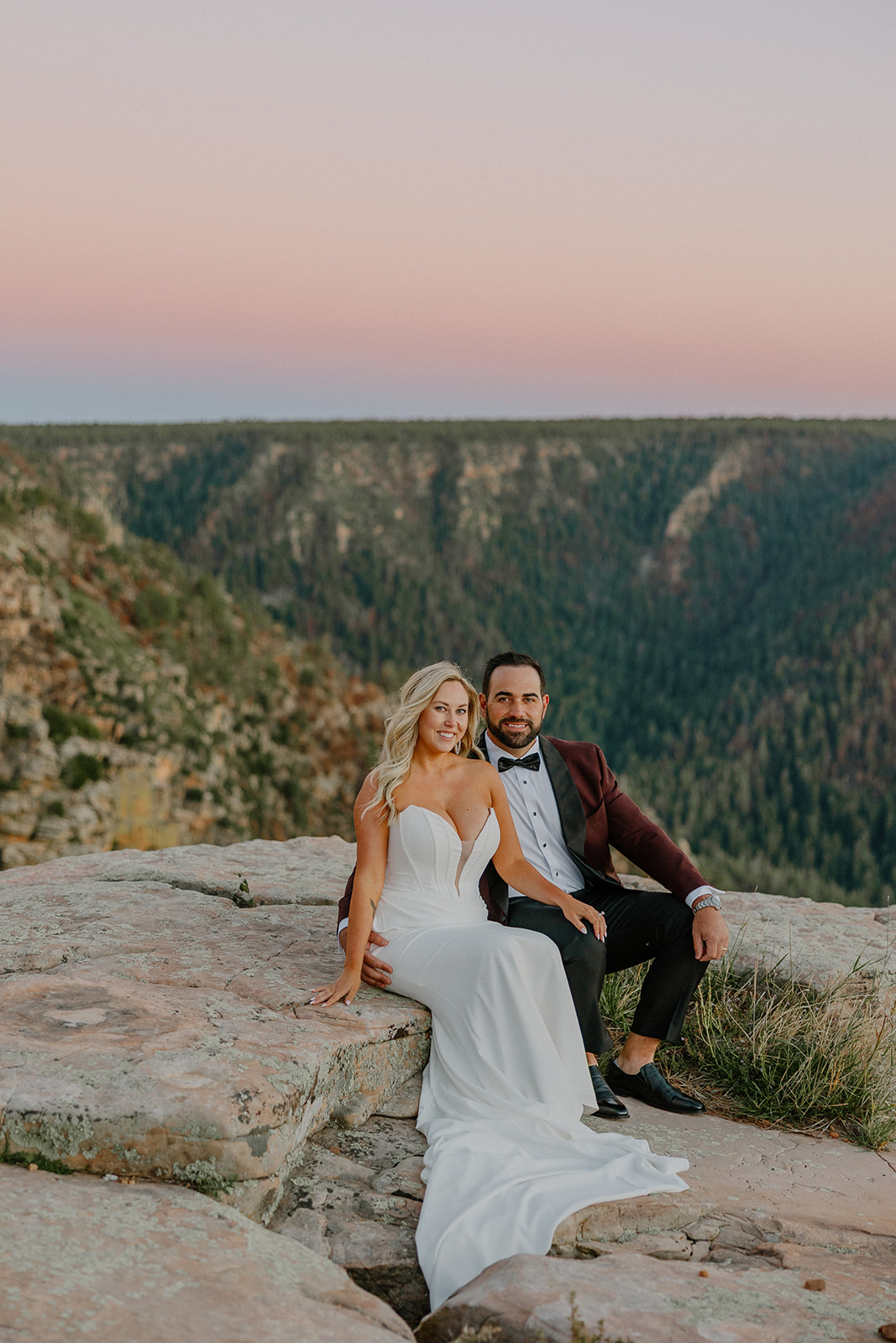 Bride and groom seated together on a rocky overlook with sweeping canyon views during sunset portraits.