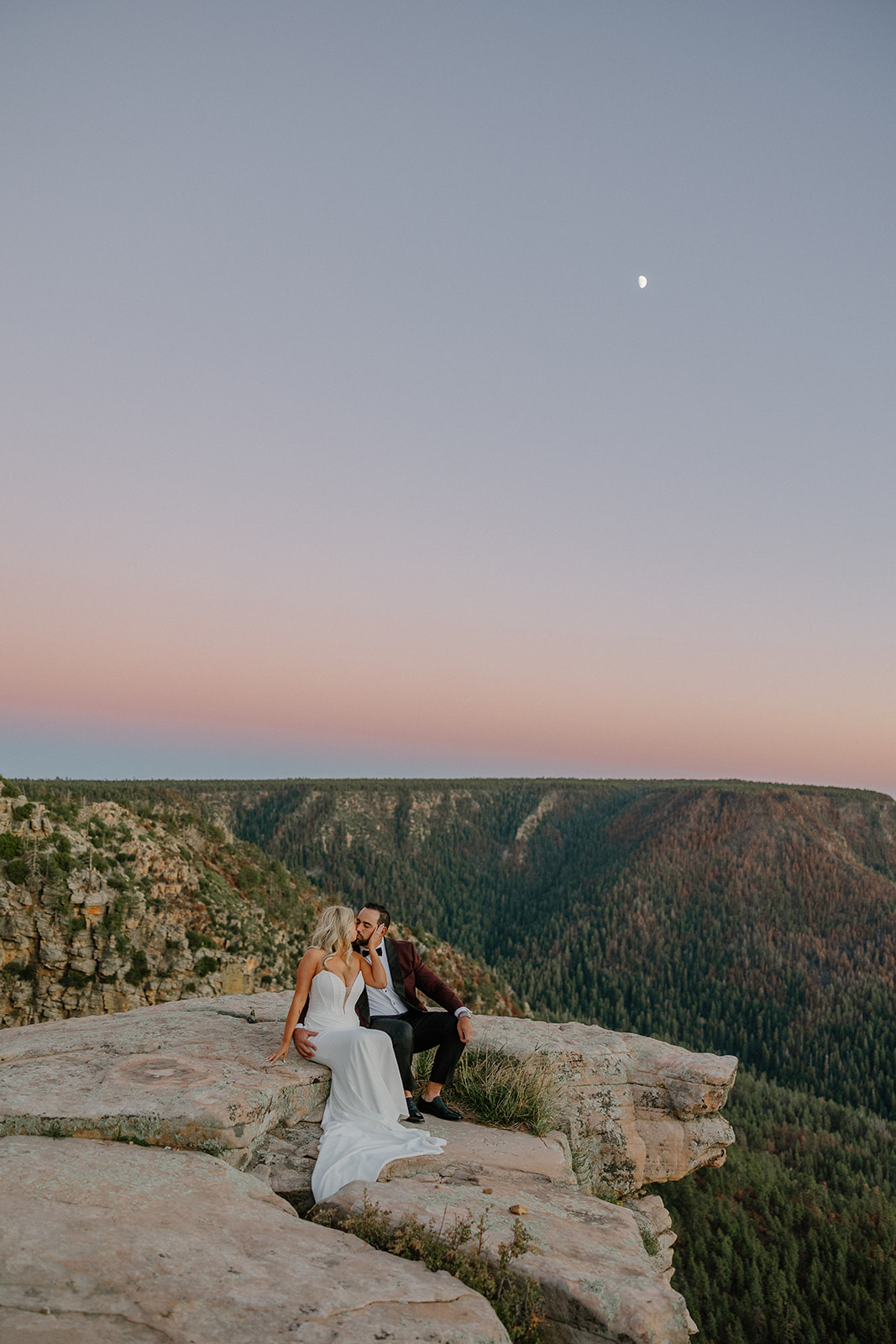 Bride and groom sit together on a cliff edge at sunset, overlooking a forested canyon during intimate wedding portraits.