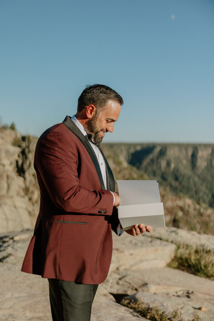 Groom reads a handwritten letter while standing on a cliffside during an intimate wedding moment.