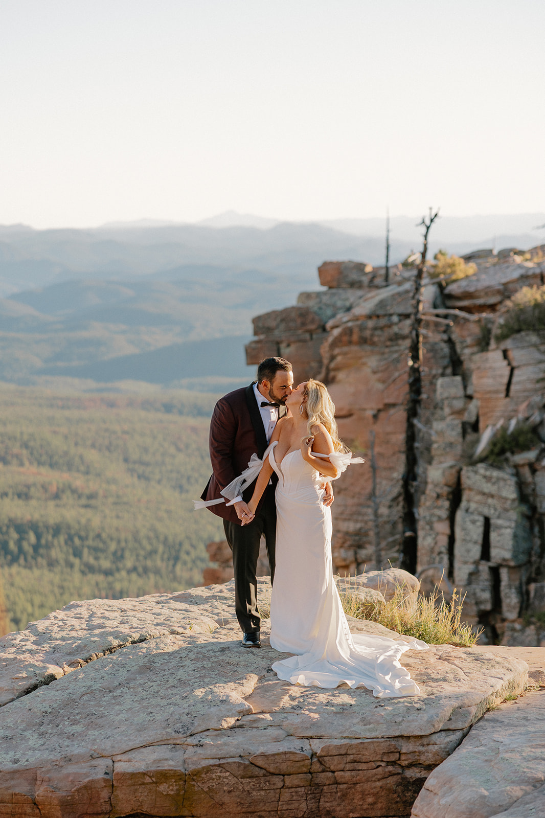 Bride and groom stand together on a cliffside ledge overlooking forested mountains during intimate sunset portraits.