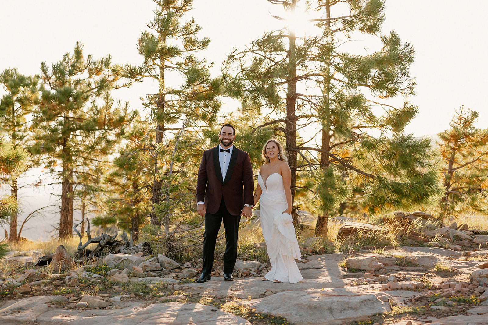 Bride and groom stand holding hands among pine trees during golden hour portraits at a Payson wedding venue.
