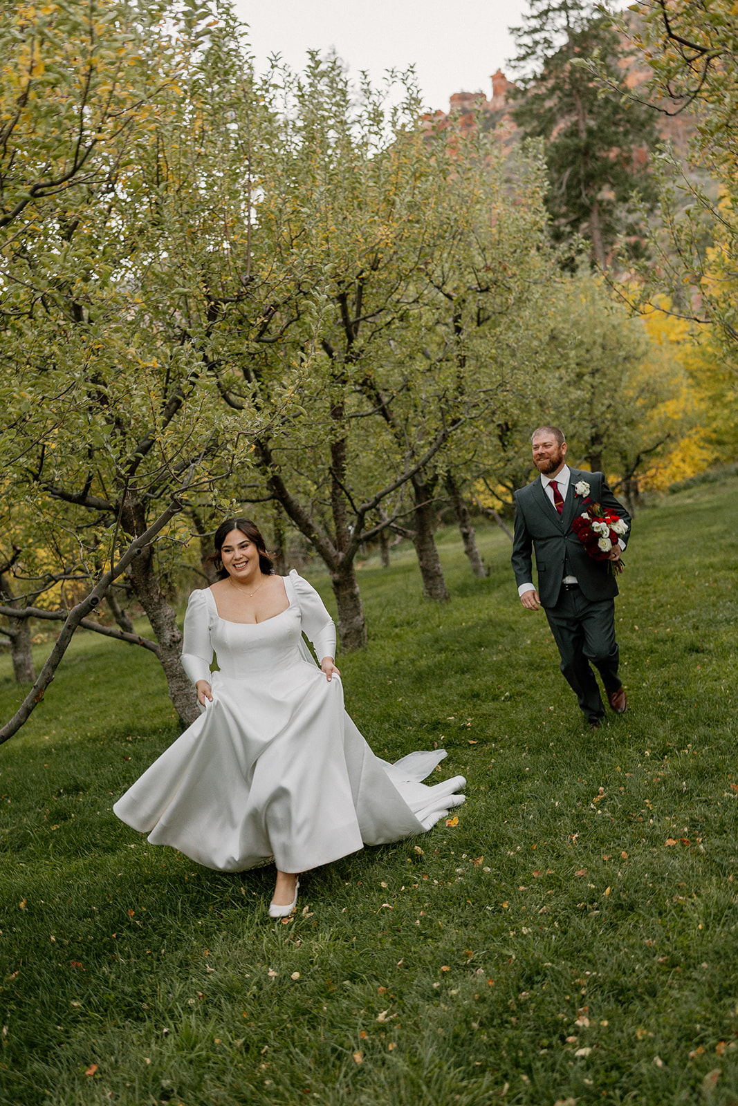 Bride running through an orchard with her partner following during golden hour portraits