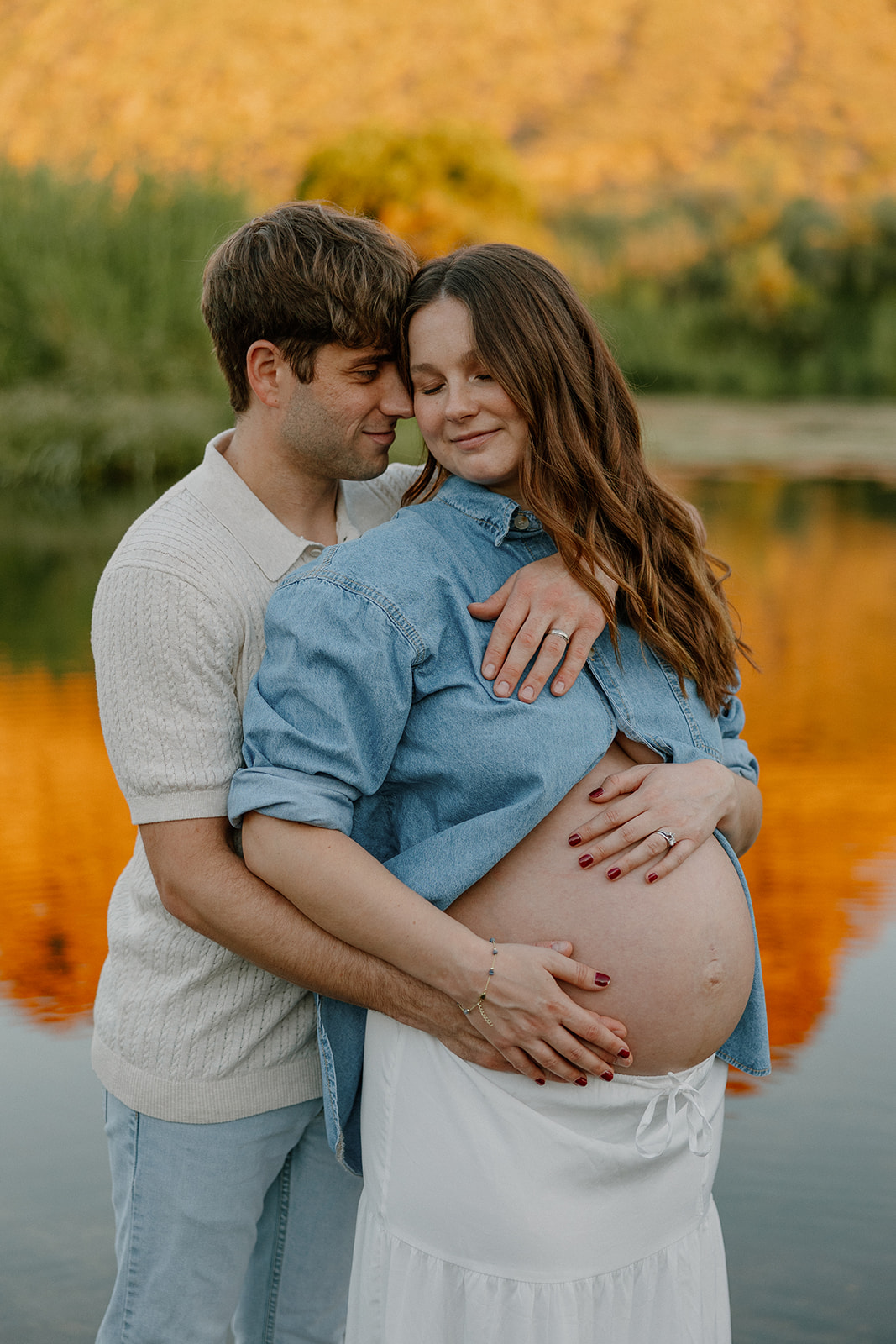 Close-up of couple embracing with hands on baby bump and warm sunset tones in the background
