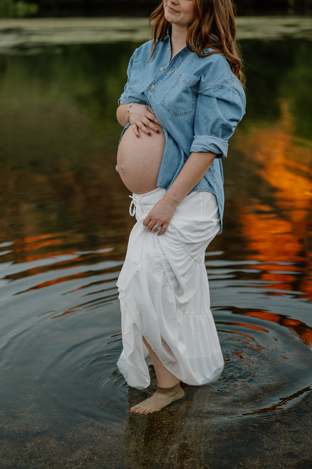 Close-up of baby bump and flowing skirt in shallow river water with warm reflections
