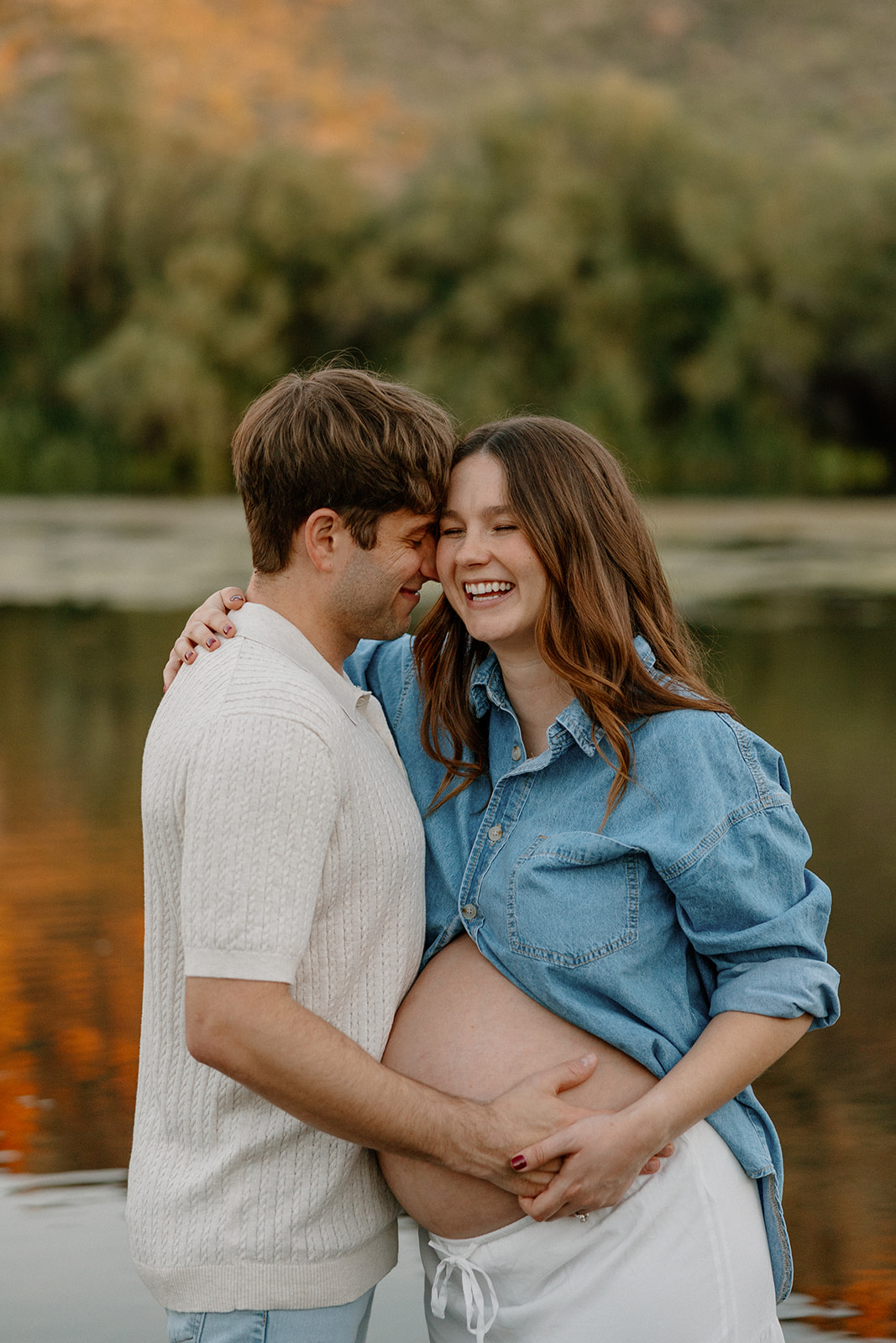 Close-up of expecting couple smiling together with hands on baby bump by the water
