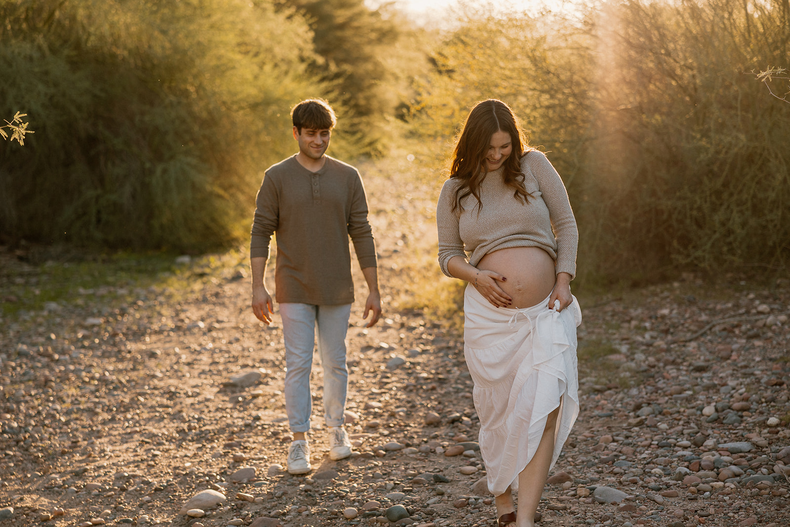 Pregnant woman walking along a rocky desert path during sunset with partner nearby, Arizona maternity photographer
