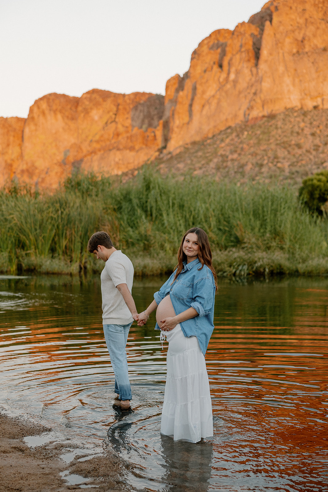 Pregnant woman standing in the river while partner sits nearby during a relaxed Arizona maternity session, Arizona maternity photographer
