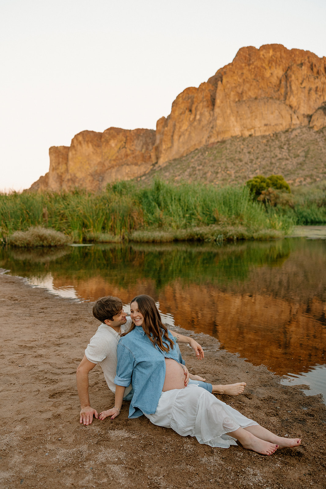 Couple sitting together on the riverbank with soft sunset light and mountain backdrop, Arizona maternity photographer
