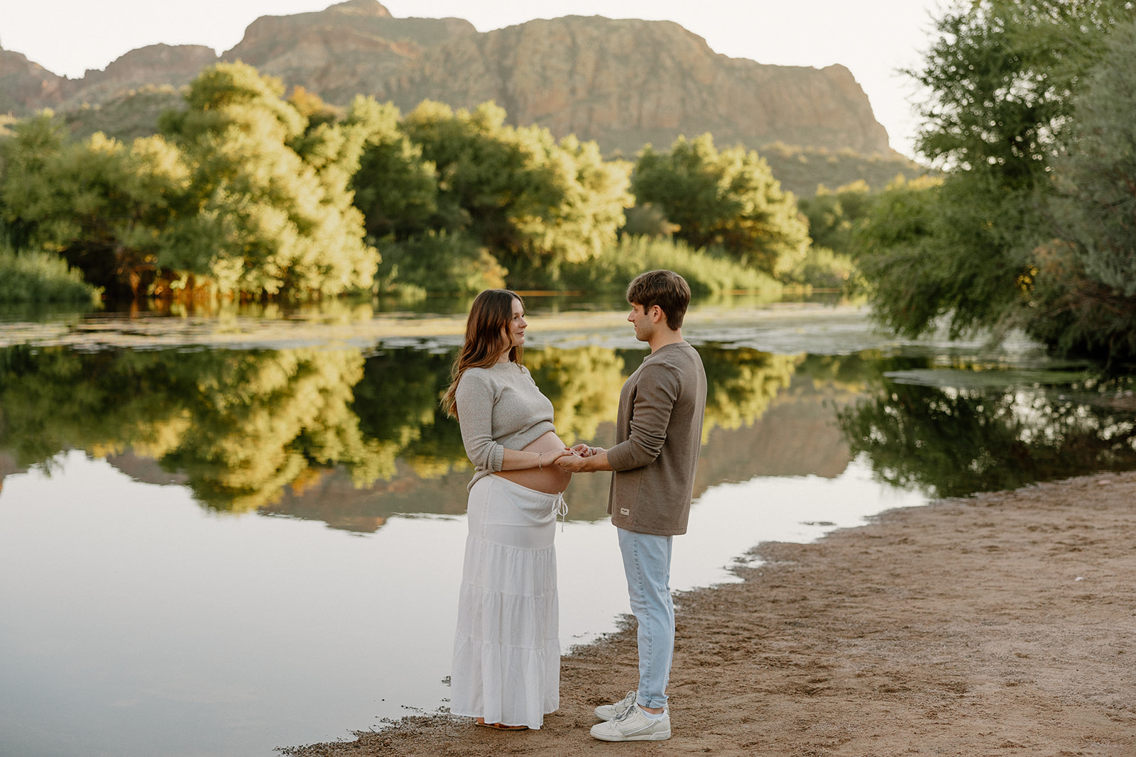 Expecting couple holding hands along the riverbank with soft evening light in Phoenix
