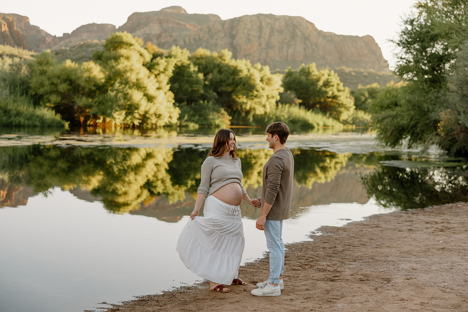 Expecting couple holding hands along the riverbank with soft evening light and calm water
