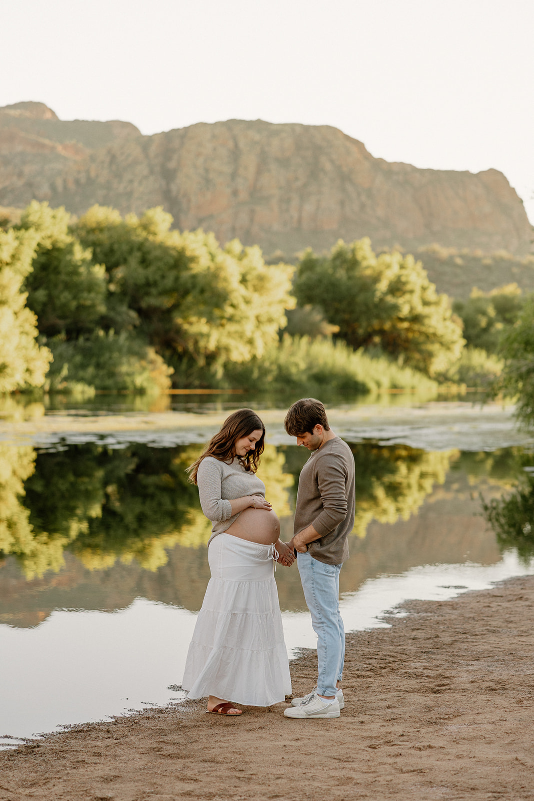Expecting couple standing close together by the river with mountains in the background, Arizona maternity photographer