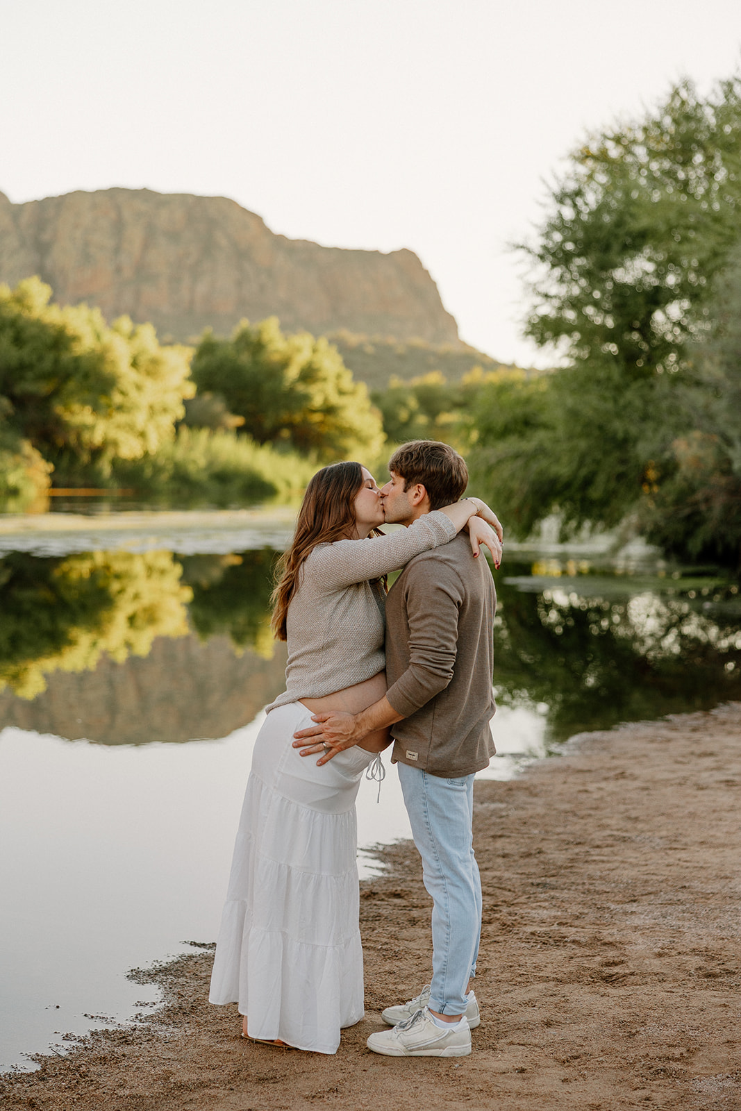 Couple kissing by the riverbank with soft evening light and calm water reflections, Arizona maternity photographer
