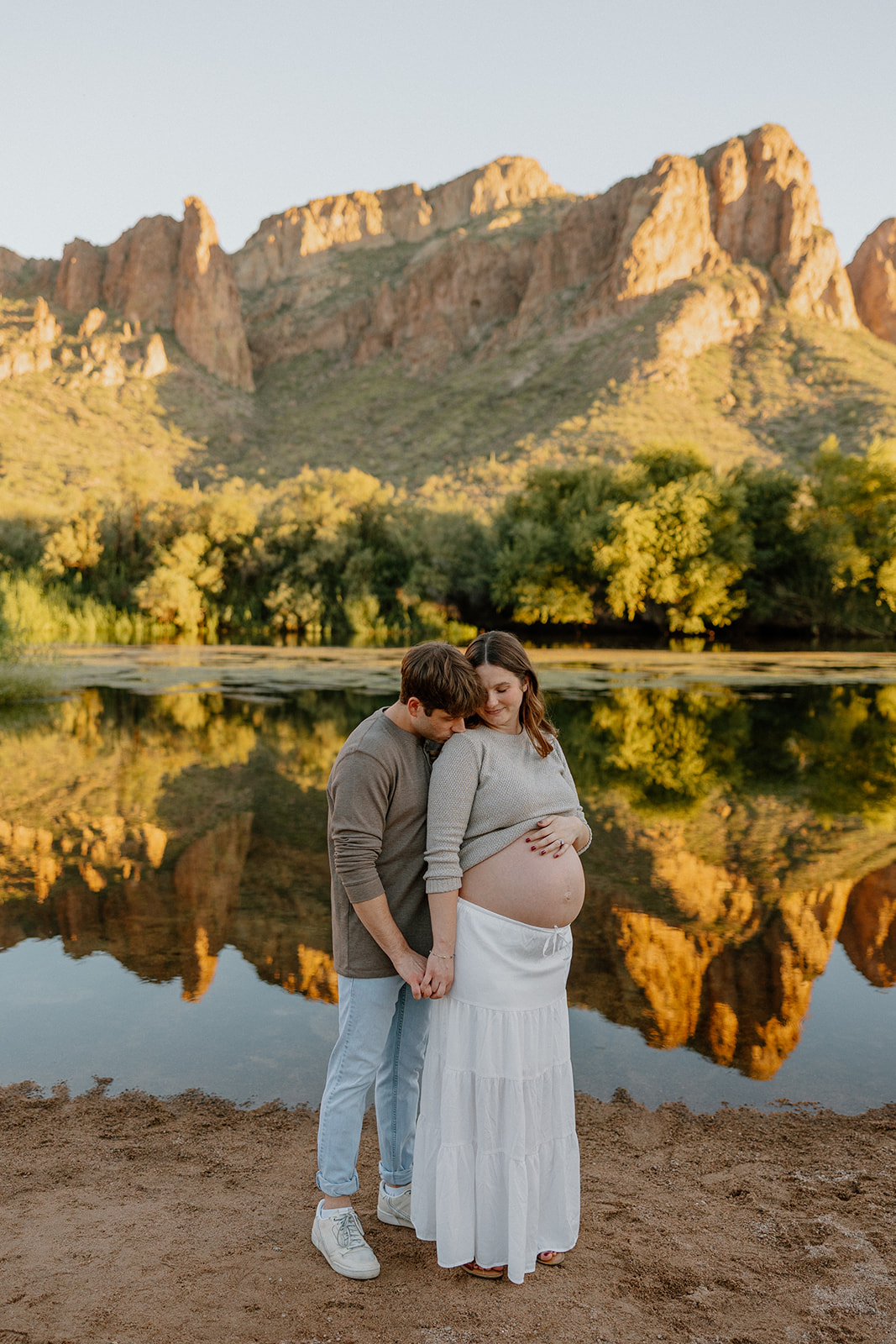Couple holding each other by the Salt River with mountain reflections at sunset, Arizona maternity photographer
