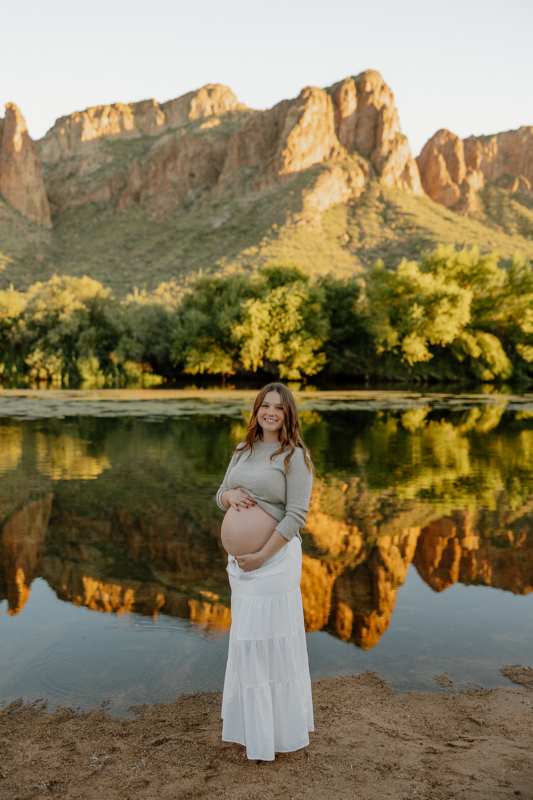 Expecting mother standing along the Salt River with mountains reflecting in the water at sunset
