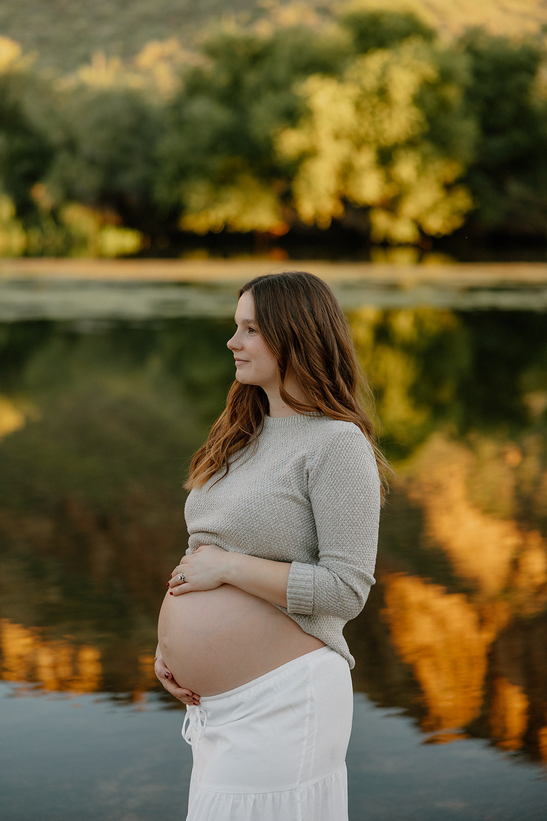 Pregnant woman standing by the river with soft sunset reflections behind her, Arizona maternity photographer
