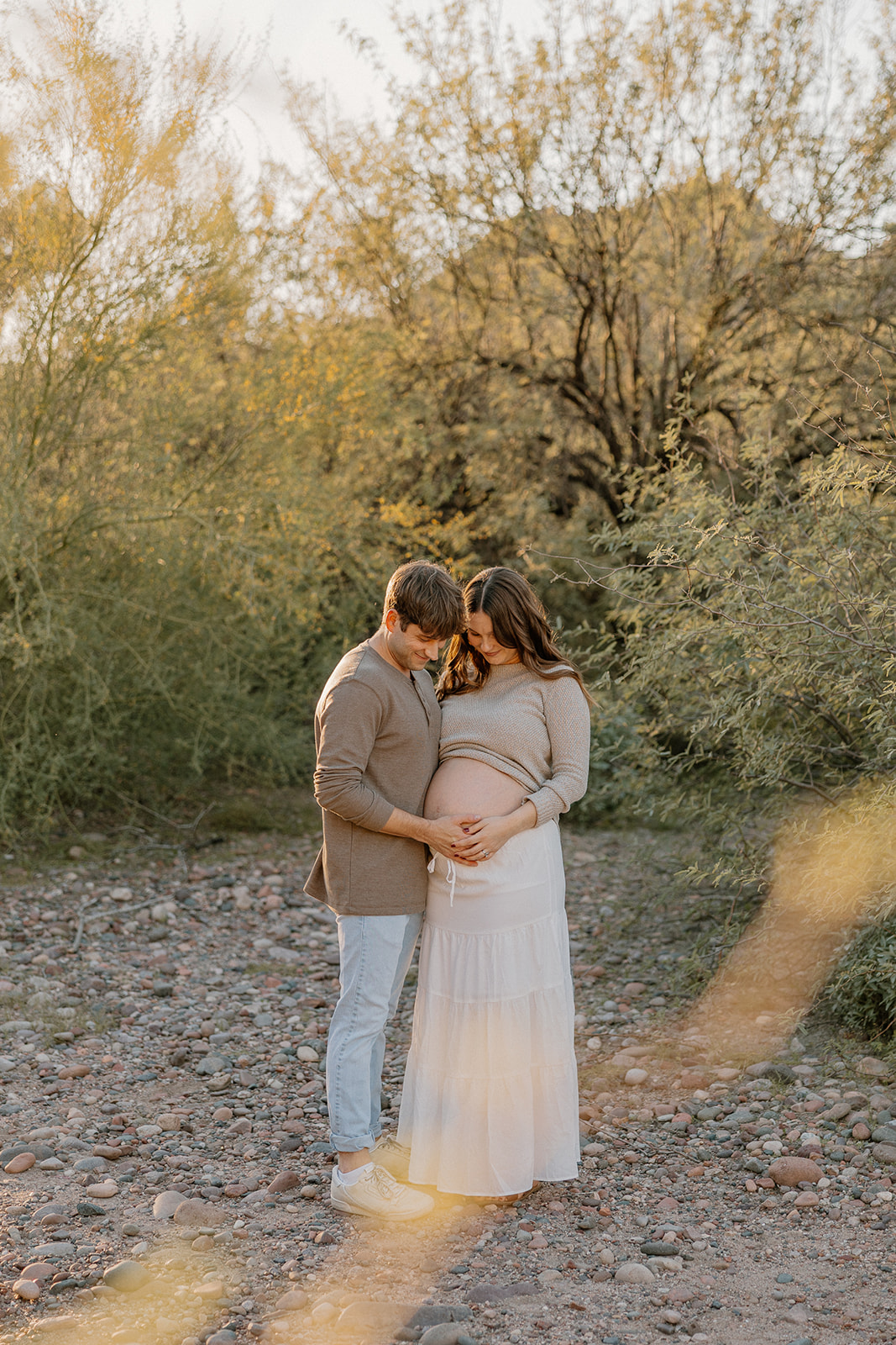 Couple standing close together on a desert trail, hands on baby bump during golden hour, Arizona maternity photographer